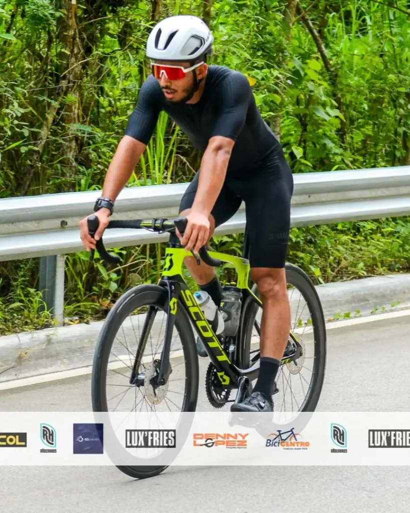 Deportista en bicicleta de ruta en un entorno natural con vegetación y camino pavimentado.