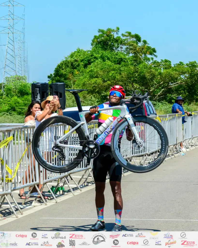 Ciclista con casco y gafas sosteniendo su bicicleta en una competencia al aire libre.