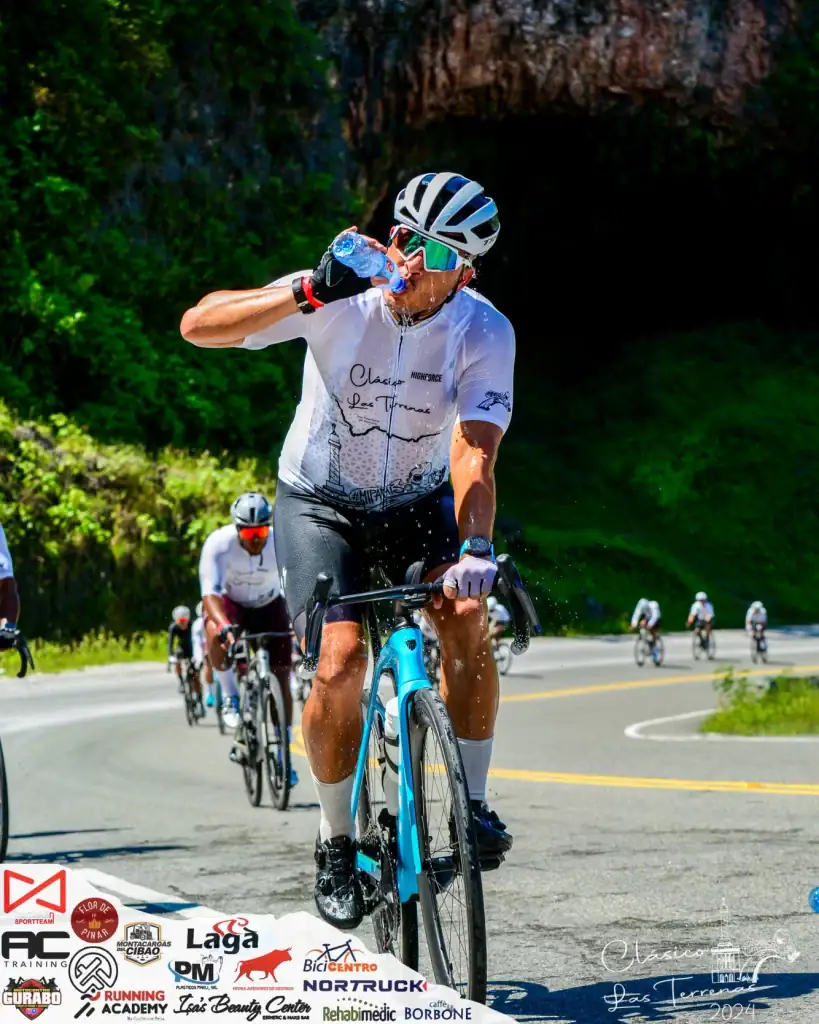 Ciclista en ruta de montaña bebiendo agua durante entrenamiento en un día soleado.
