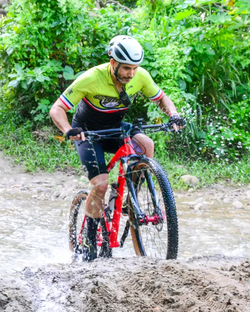 Aventurero en bicicleta de montaña atravesando un río con vegetación verde.