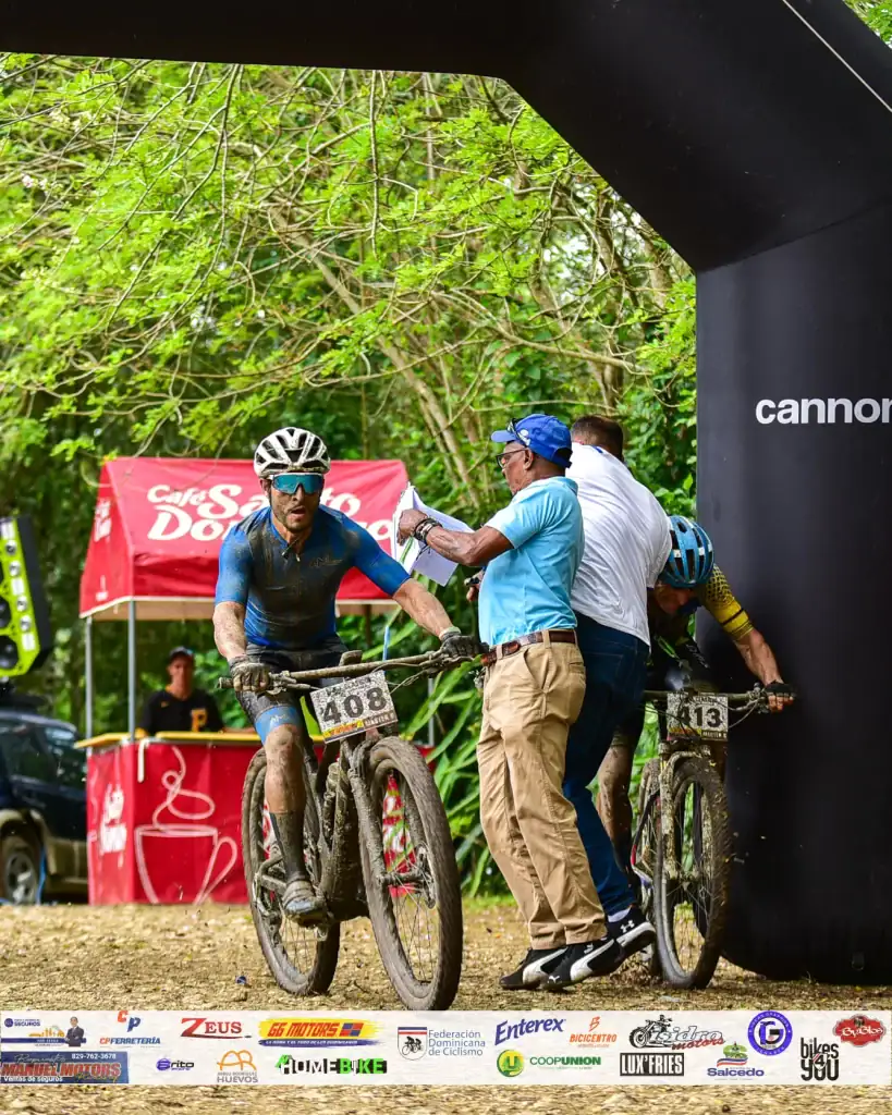 Ciclista en carrera de mountain bike en un entorno natural con árboles verdes y un arco de meta negr.