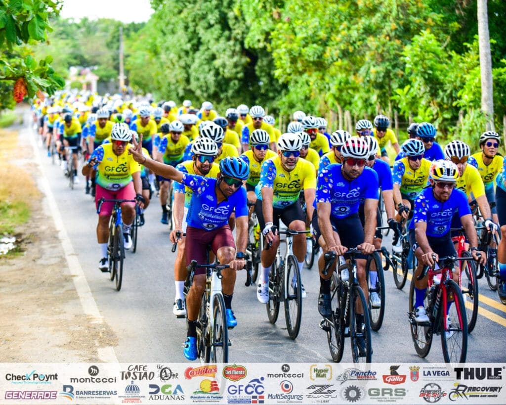 Ciclistas en una competencia de ciclismo en un camino rodeado de árboles verdes.