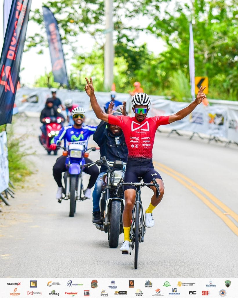 Denny Lopez celebrando en una carrera en bicicleta con apoyo de motocicletas y espectadores.