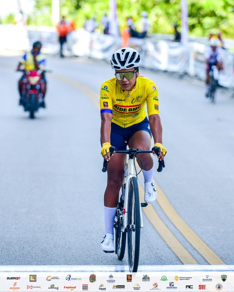 Ciclista profesional Denny Lopez en competencia, vestido con uniforme amarillo y casco, pedaleando e.