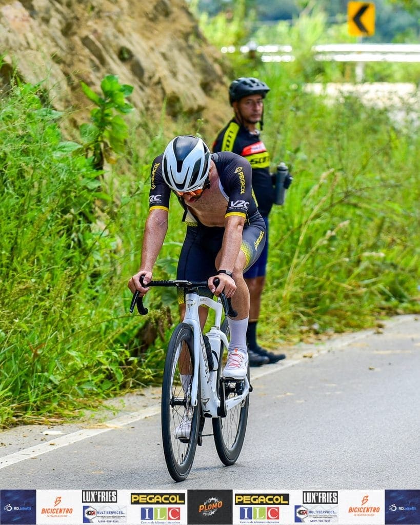 Ciclista en ruta de montaña con casco, ropa deportiva y bicicleta en un camino rodeado de vegetación.